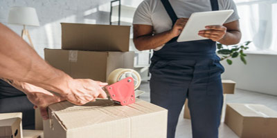A man filling out a form on a clip board and a pair of hands taping up a box with packed boxes around them.
